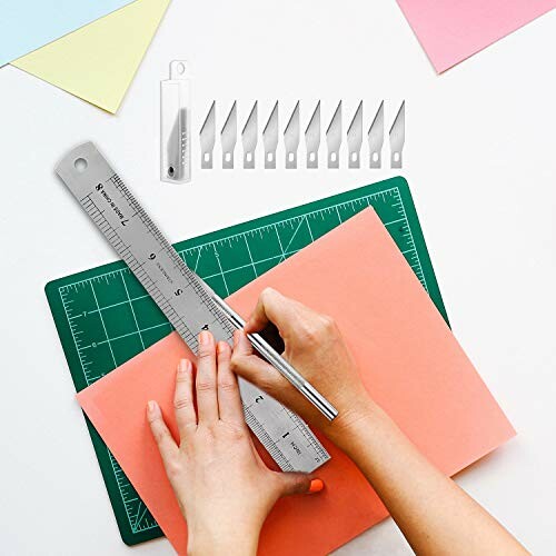 Hands using steel ruler and craft knife on colored paper with extra blades and cutting mat.